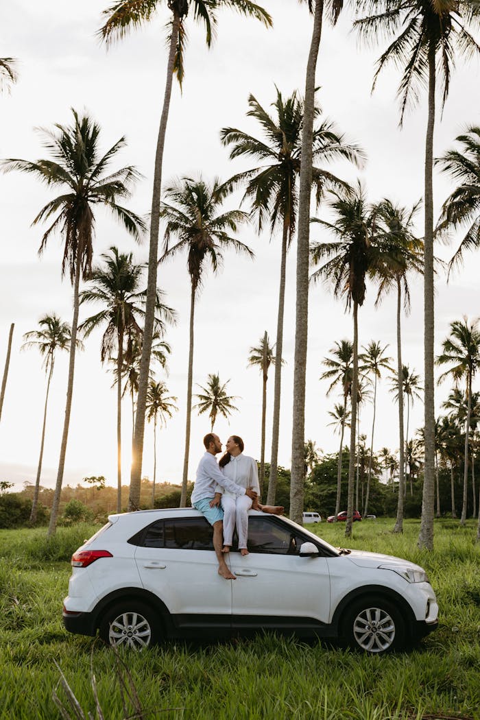 A couple enjoys a romantic moment on an SUV amidst tropical palm trees during sunset.