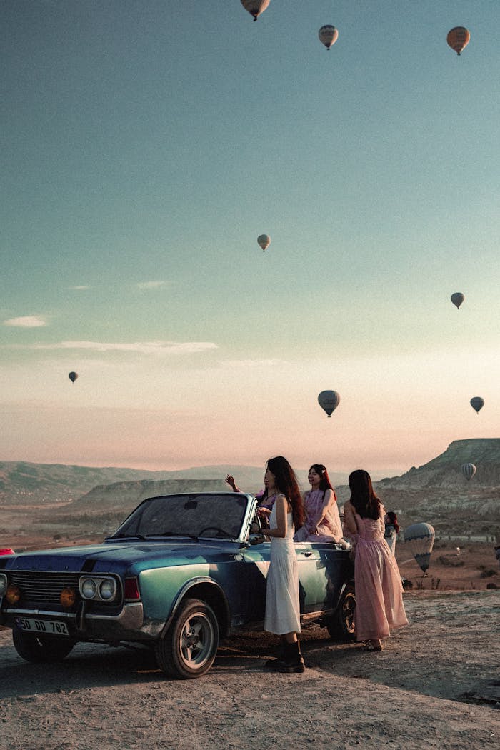 Hot air balloons fill the sky over Cappadocia, Turkey, with people enjoying the view from a vintage car.