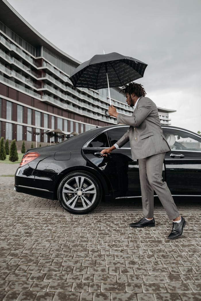 Man in a gray suit holding an umbrella while opening a black car door outside a modern building.